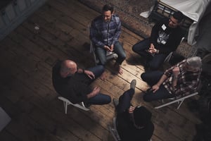 Five men form a Men's Group, seated in a circle on wooden chairs, engaged in a discussion. The room's wooden floor complements the intimate setting, with part of a bed visible in the background, symbolizing their deep connection and male friendship.