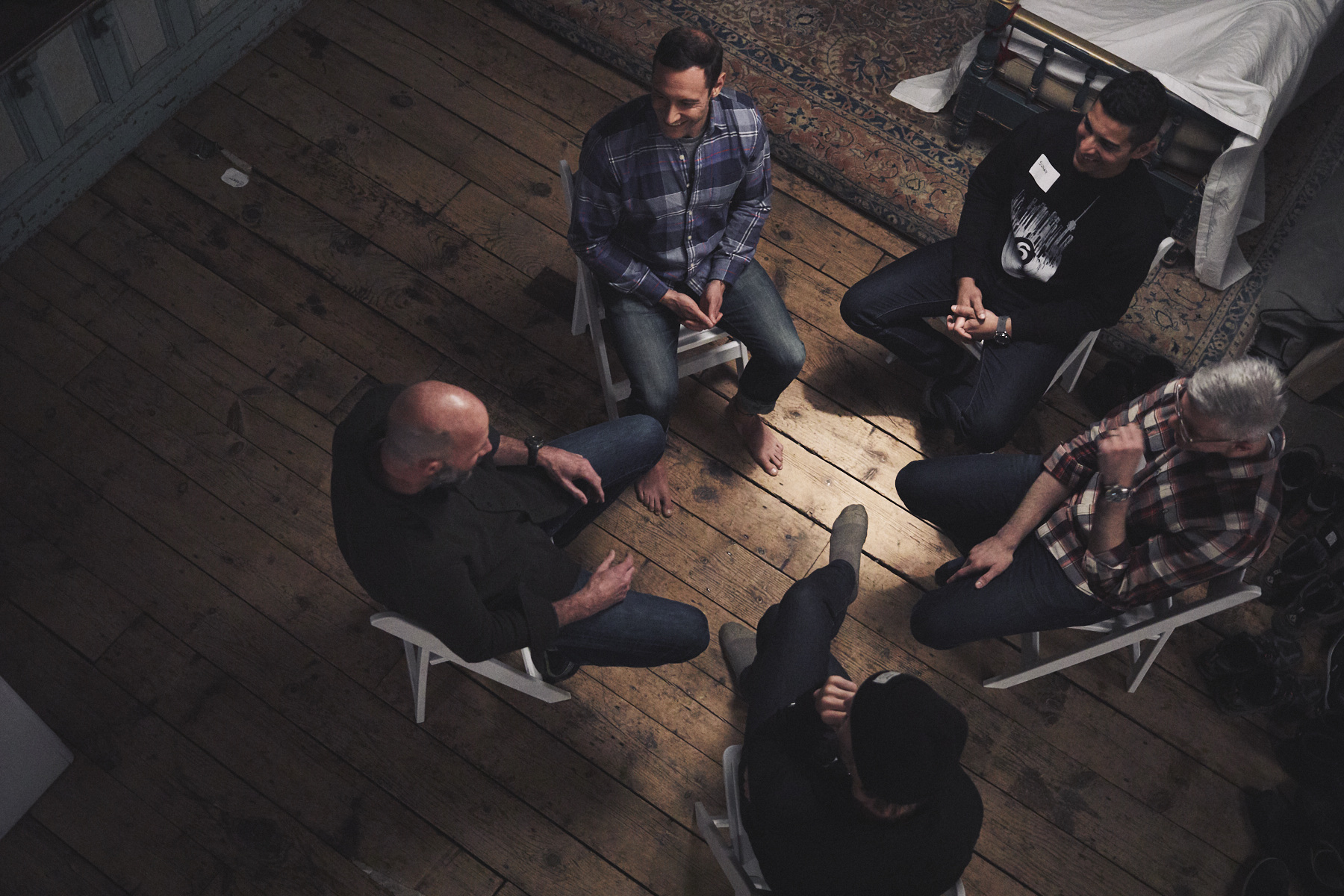 Five men form a Men's Group, seated in a circle on wooden chairs, engaged in a discussion. The room's wooden floor complements the intimate setting, with part of a bed visible in the background, symbolizing their deep connection and male friendship.